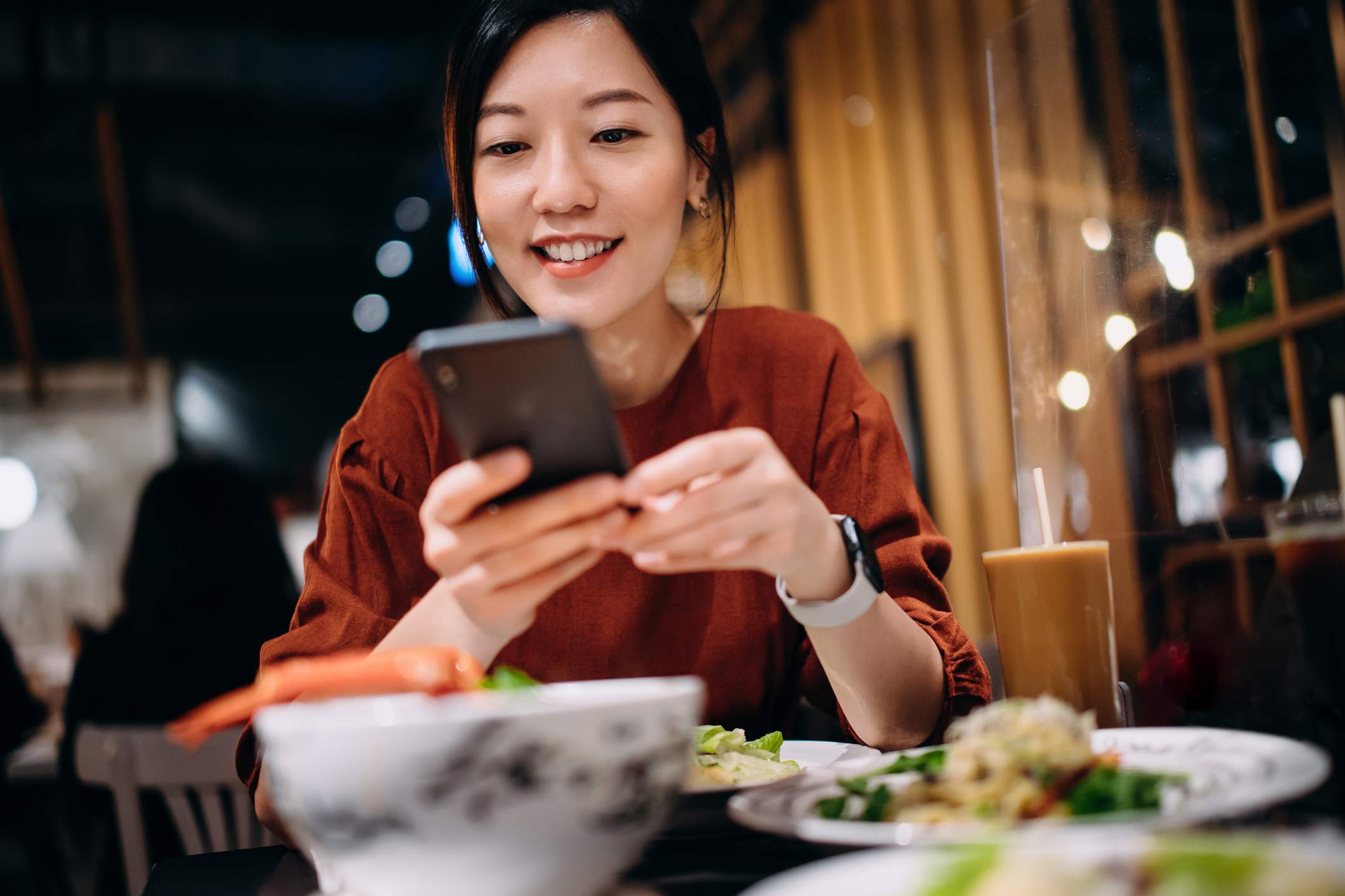Image depicts a person at a restaurant using their phone. They are wearing a red shirt and a smartwatch and are smiling down at their food and their phone.